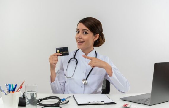 impressed-young-female-doctor-wearing-medical-robe-stethoscope-sitting-desk-with-medical-tools-laptop-holding-pointing-credit-card-isolated_141793-68735